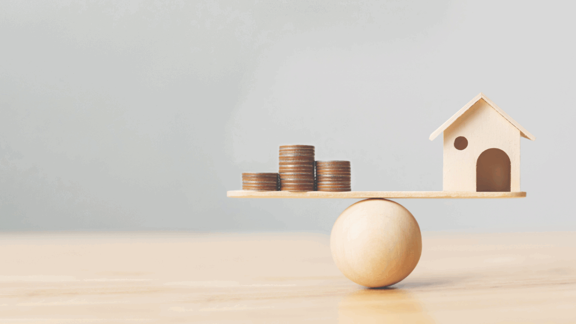 Stacks of coins and a small wooden house balanced on a wooden plank atop a round wooden ball