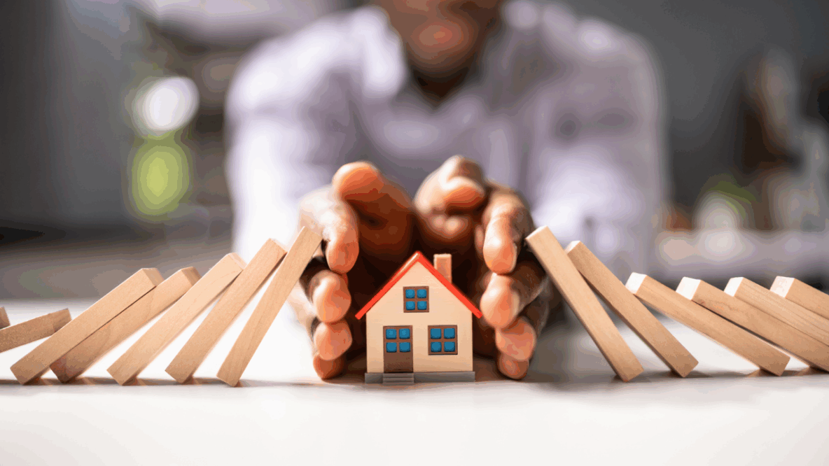 Person's hands protecting a small model house from falling dominoes, symbolising home protection and insurance